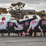 drift cars in the background. six girls embracing each other in a line wearing hoodies