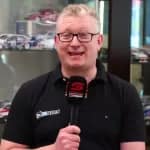 Aaron Noon with a v8 Sleuth logo holding a microphone with supercars talking to camera